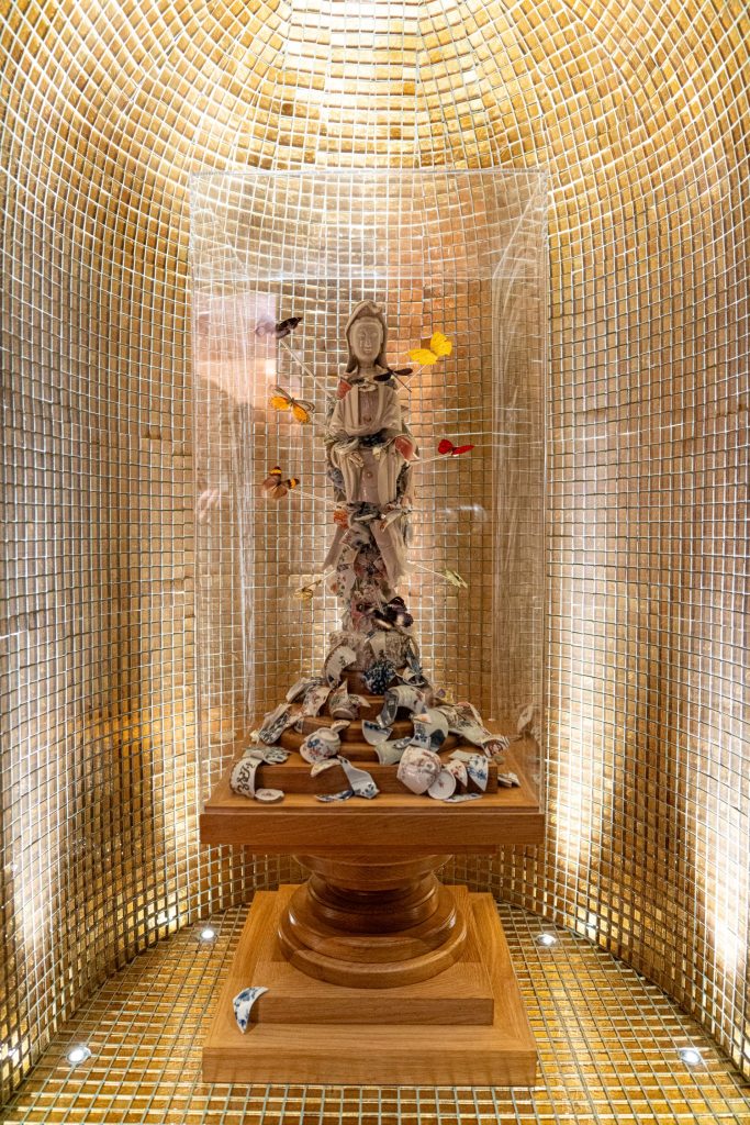 The White Lady with Dragonfly (2025). Broken porcelain fragments, Guan Yin sculpture, glass vitrine. Courtesy of Adrian Sassoon Gallery.